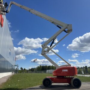 Bomlift Nifty HR28 lyfter arbetare upp mot en byggnadsvägg under en klarblå himmel med spridda moln. I bakgrunden syns flaggor och träd, och marken är täckt av gräs.